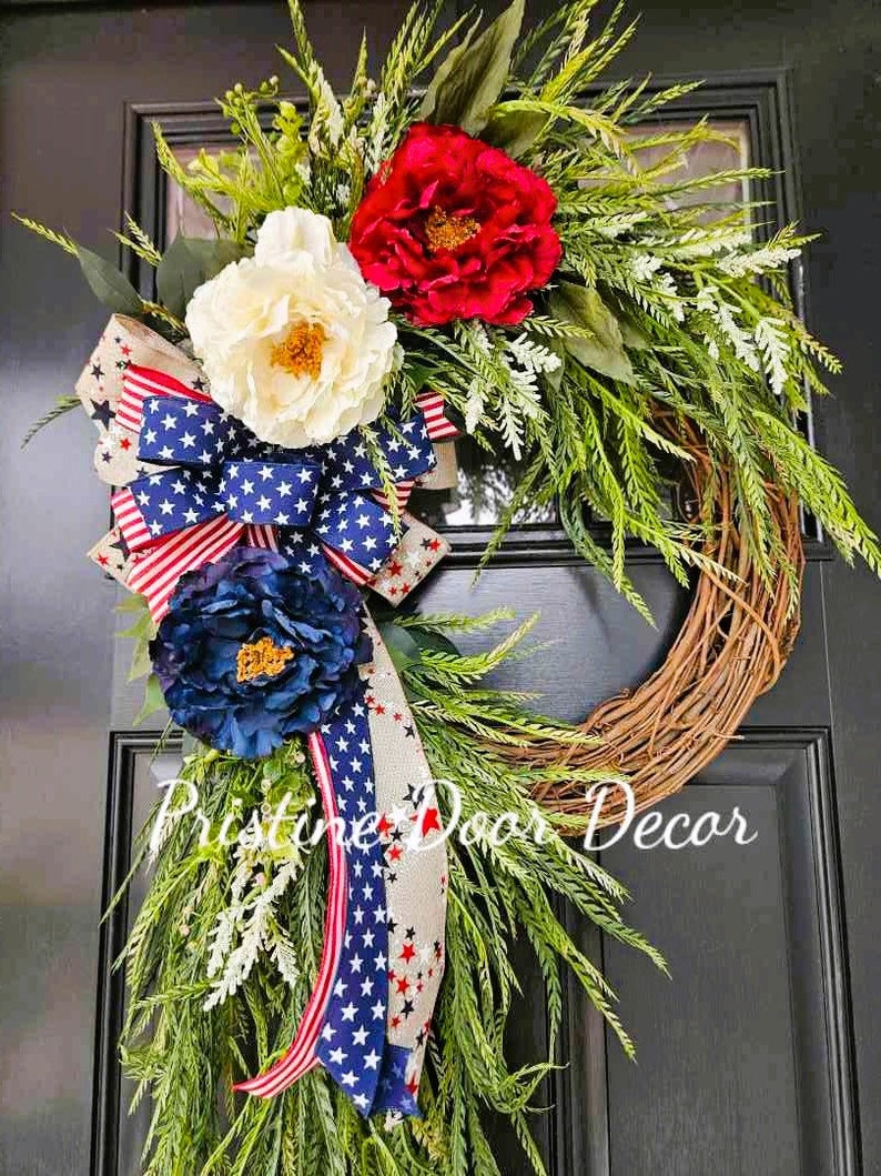 May include: A patriotic wreath with red, white, and blue flowers and a ribbon with stars and stripes. The wreath is made of grapevine and is decorated with greenery. The text "Pristine Door Decor" is visible in the bottom right corner of the image.