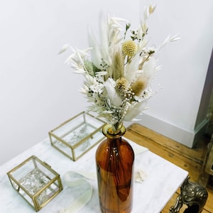 Op de afbeelding: Een boeket gedroogde witte bloemen met groene accenten in een bruine glazen vaas op een witte marmeren tafel met gouden accenten.