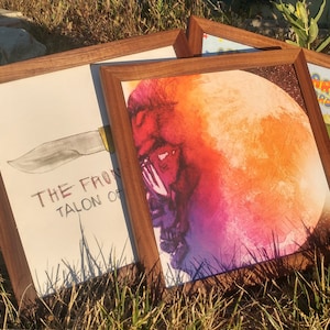 May include: Three framed art prints with dark wood frames are displayed outdoors. One print features a knife illustration with the text "THE FRONT TALON OF". Another print shows a colorful, surreal image of a face and a moon. The third print has a cartoon image.