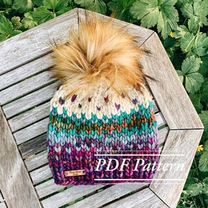 Peut inclure: Un bonnet tricoté coloré avec un pompon moelleux. Le bonnet a une base blanche avec des rayures violettes, turquoise et brunes. Le bonnet est posé sur une surface en bois. PDF Pattern.
