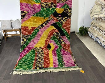 Puede incluir: Una vibrante alfombra rectangular con un diseño abstracto y colorido. La alfombra presenta una mezcla de formas geométricas rosas, amarillas, verdes, rojas y negras. Tiene un borde con flecos y se exhibe contra una pared blanca.