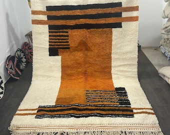 May include: A large, rectangular rug with a cream-colored background and geometric patterns in shades of orange, black, and brown. The rug has a thick, textured pile and a fringe along the bottom edge. The design features horizontal and vertical lines.