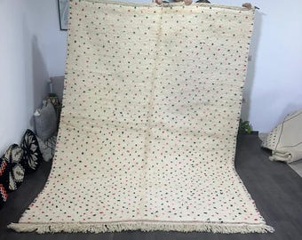May include: A white Moroccan rug with a scattered pattern of red, green, and blue dots. The rug has a fringe border and is being held up by two people.