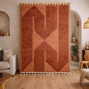 Peut inclure: Un grand tapis rectangulaire de couleur rouille avec un motif g&eacute;om&eacute;trique accroch&eacute; au mur. Le tapis pr&eacute;sente un motif textur&eacute; avec une forme de losange centrale et des rayures verticales. Des glands cr&egrave;me ornent les bords sup&eacute;rieur et inf&eacute;rieur. Le tapis est un &eacute;l&eacute;ment d&eacute;coratif.