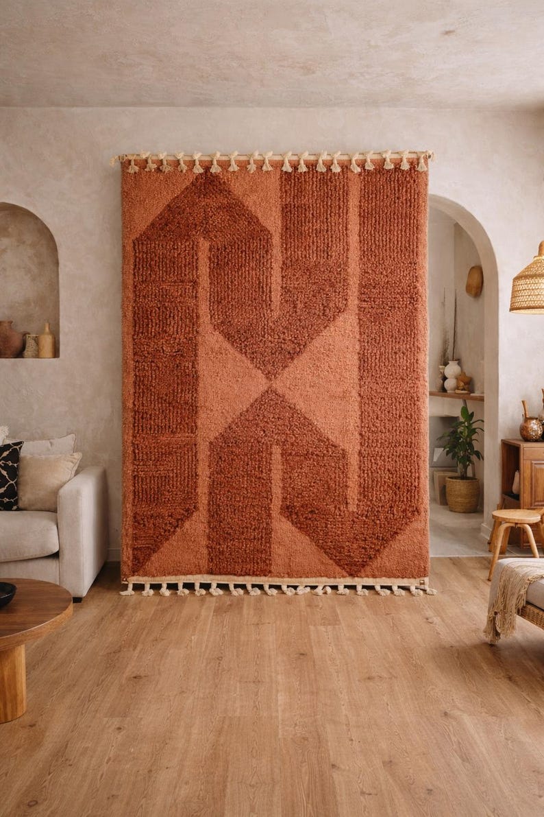 Peut inclure: Un grand tapis rectangulaire de couleur rouille avec un motif g&eacute;om&eacute;trique accroch&eacute; au mur. Le tapis pr&eacute;sente un motif textur&eacute; avec une forme de losange centrale et des rayures verticales. Des glands cr&egrave;me ornent les bords sup&eacute;rieur et inf&eacute;rieur. Le tapis est un &eacute;l&eacute;ment d&eacute;coratif.