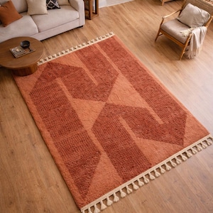 Peut inclure: Un tapis &agrave; motifs g&eacute;om&eacute;triques orange et p&ecirc;che avec une frange cr&egrave;me. Le tapis est rectangulaire et pr&eacute;sente un motif r&eacute;p&eacute;titif de formes angulaires. Le tapis est pos&eacute; sur un parquet dans un salon.