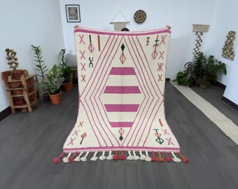 Op de afbeelding: Een crèmekleurig tapijt met een geometrisch ontwerp in roze. Het tapijt heeft een centrale ruitvorm met horizontale strepen en is omzoomd met roze lijnen en decoratieve symbolen. Kwasten in verschillende kleuren sieren de onderrand.