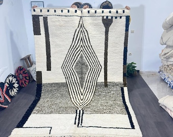 Op de afbeelding: Een groot, rechthoekig vloerkleed met een geometrisch ontwerp. Het vloerkleed heeft een crèmekleurige achtergrond met een centrale ruitvorm in zwart en bruin. Het onderste gedeelte is grijs en de randen zijn afgewerkt met franjes.