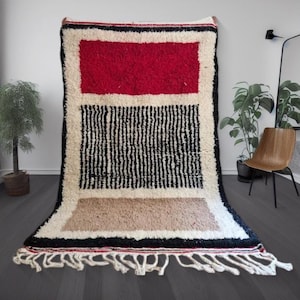 May include: A rectangular, handmade rug with a geometric design. The rug features a red rectangle at the top, a black and white striped section, and a tan rectangle at the bottom. The rug has a white border and fringe.