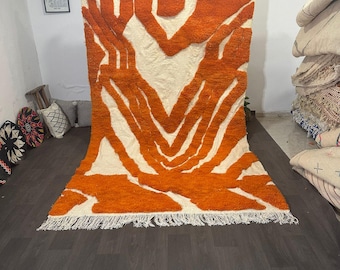May include: A large, rectangular rug with a bold, abstract design. The rug features a cream-colored background with a striking pattern of orange, vertical lines and shapes. The rug has a fringed edge and is displayed indoors.