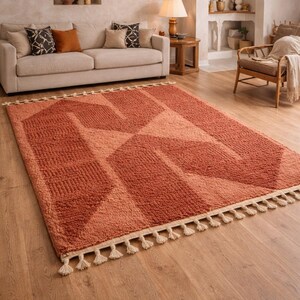 Peut inclure: Tapis rectangulaire terracotta avec un motif g&eacute;om&eacute;trique de rectangles et de triangles. Le tapis a une surface textur&eacute;e et une frange cr&egrave;me. Il est pos&eacute; sur un sol en bois dans un salon. Mesure environ 200 cm x 300 cm.
