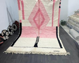 Puede incluir: Una alfombra rectangular grande con un diseño geométrico. La alfombra presenta una forma de diamante central en tonos de rosa, enmarcada por acentos blancos y negros. La alfombra tiene un borde con flecos y se muestra sobre un fondo neutro.