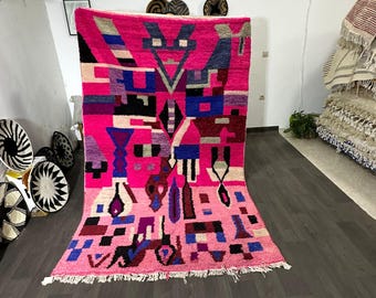 Puede incluir: Una vibrante alfombra rosa con un diseño geométrico que presenta formas azules, negras y blancas. La alfombra tiene flecos en el borde inferior y se exhibe contra una pared blanca. El diseño incluye formas y patrones abstractos.