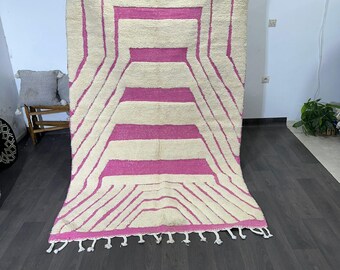 Op de afbeelding: Een groot, rechthoekig vloerkleed met een crèmekleurige achtergrond en een geometrisch ontwerp in felroze. Het ontwerp heeft horizontale strepen en schuine lijnen. Het vloerkleed heeft een franje langs de onderrand.