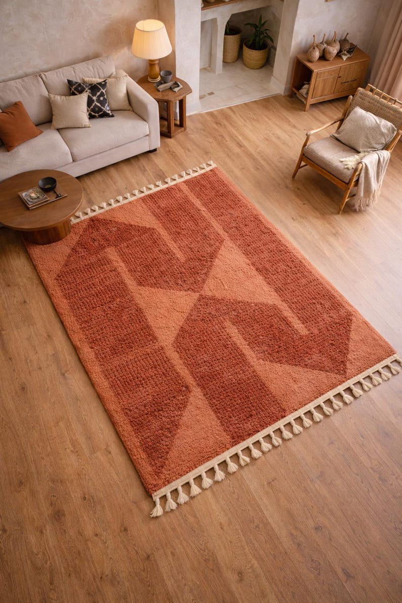 Peut inclure: Un tapis &agrave; motifs g&eacute;om&eacute;triques orange et p&ecirc;che avec une frange cr&egrave;me. Le tapis est rectangulaire et pr&eacute;sente un motif r&eacute;p&eacute;titif de formes angulaires. Le tapis est pos&eacute; sur un parquet dans un salon.