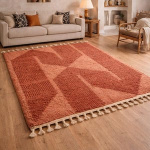 Peut inclure: Tapis rectangulaire de couleur terre cuite avec un motif g&eacute;om&eacute;trique. Le tapis pr&eacute;sente une surface textur&eacute;e et une frange cr&egrave;me. Il est pos&eacute; sur un sol en bois, cr&eacute;ant une ambiance chaleureuse dans un salon.
