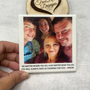 Puede incluir: Un posavasos de cerámica blanca con una foto de una familia de cuatro. La foto muestra un hombre, una mujer y dos niños. El texto debajo de la foto dice: "NO MATTER WHERE YOU GO, & NO MATTER WHAT YOU DO, YOU WILL ALWAYS HAVE US CHEERING FOR YOU! - MOM".