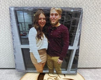 May include: A slate coaster with a photo of a couple, a man and a woman, smiling at the camera. The coaster is sitting on a wooden round with the text "True Love Designs" burned into the wood.