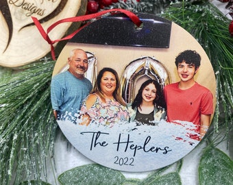 May include: A round, wooden Christmas ornament with a photo of a family of four. The ornament is white with a blue and white paint splatter design. The text "The Heplers 2022" is printed on the ornament.