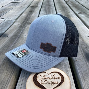 May include: A gray and black trucker hat with a brown leather patch that says "STATE" on it. The hat is sitting on a wooden slice with the words "True Love Designs" burned into it.