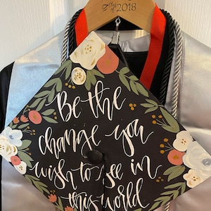 May include: A black graduation cap with the quote "Be the change you wish to see in this world" in white script, adorned with floral accents in pink, white, and green. The cap is displayed on a wooden hanger with the name "Tammy" and "Class of 2018".
