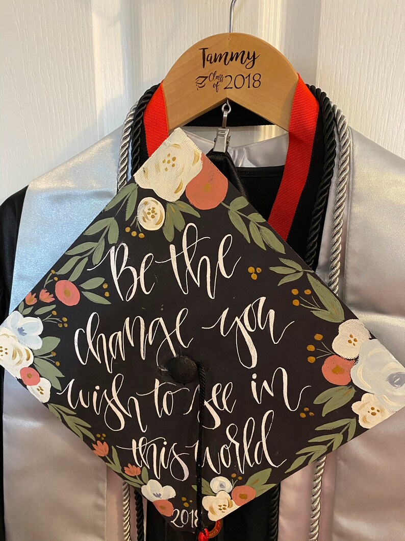 May include: A black graduation cap with the quote "Be the change you wish to see in this world" in white script, adorned with floral accents in pink, white, and green. The cap is displayed on a wooden hanger with the name "Tammy" and "Class of 2018".