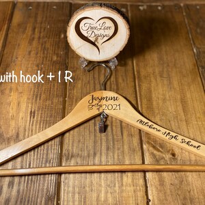 May include: A wooden clothes hanger with the name "Jasmine" and "Class of 2021" engraved on it, along with "Attleboro High School." A small charm hangs from the hook. A wooden disc with a heart design is above the hanger.