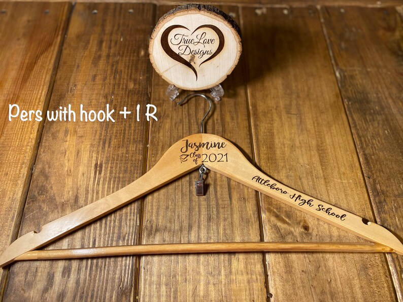 May include: A wooden clothes hanger with the name "Jasmine" and "Class of 2021" engraved on it, along with "Attleboro High School." A small charm hangs from the hook. A wooden disc with a heart design is above the hanger.