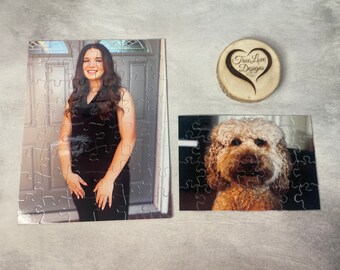 May include: Two jigsaw puzzles, one with a portrait of a woman in a black dress and the other with a portrait of a brown and white dog. The puzzles are on a gray surface with a wooden heart-shaped sign that says "True Love Designs".