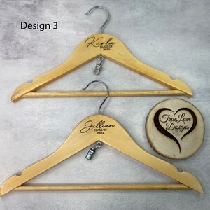 May include: Two wooden clothes hangers with metal hooks. The hangers are engraved with names and the text "CLASS OF 2022". Each hanger has a small metal clip. A wooden coaster with a heart design and the text "True Love Designs" is also visible.