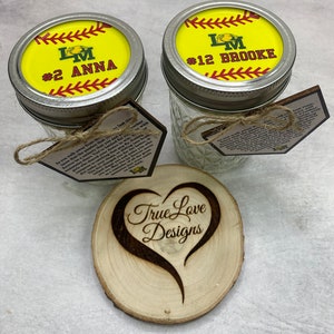 Puede incluir: Dos tarros de cristal Mason con tapas con tem&aacute;tica de softbol, etiquetados con nombres y n&uacute;meros. Tambi&eacute;n hay un posavasos de madera con un dise&ntilde;o de coraz&oacute;n y el texto "True Love Designs". Cada tarro tiene una etiqueta con texto.
