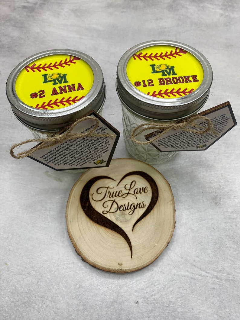 Puede incluir: Dos tarros de cristal Mason con tapas con tem&aacute;tica de softbol, etiquetados con nombres y n&uacute;meros. Tambi&eacute;n hay un posavasos de madera con un dise&ntilde;o de coraz&oacute;n y el texto "True Love Designs". Cada tarro tiene una etiqueta con texto.
