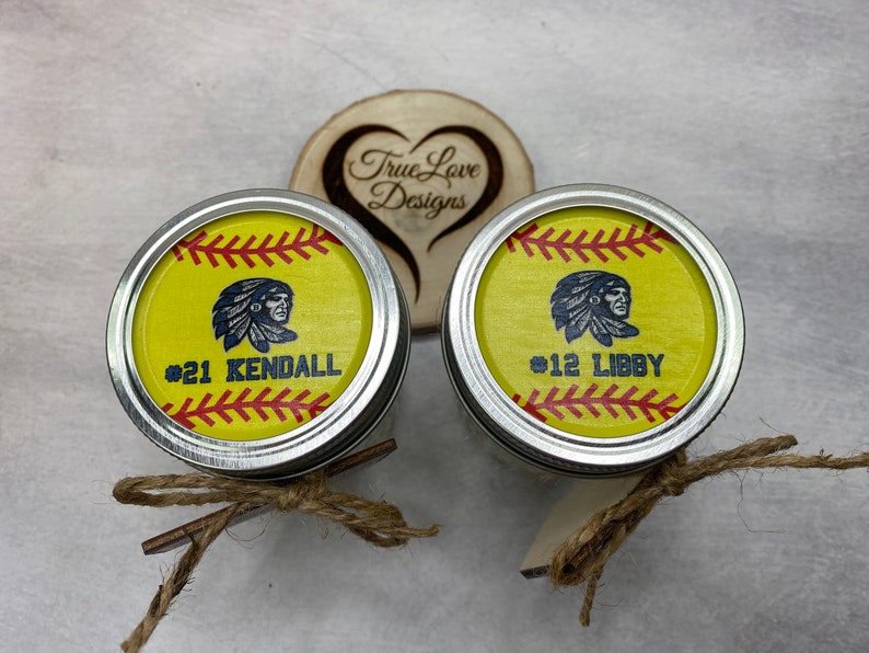 Puede incluir: Dos tapas de tarro Mason personalizadas con tem&aacute;tica de softbol con bordes plateados. Las tapas presentan un fondo amarillo con costuras rojas, un gr&aacute;fico nativo americano y los nombres "KENDALL" y "LIBBY" con sus n&uacute;meros. Atadas con hilo.