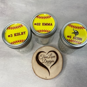 Puede incluir: Tres tarros de vidrio con tapas dise&ntilde;adas para parecerse a pelotas de softbol. Cada tapa tiene un fondo amarillo con costuras rojas y el nombre y n&uacute;mero de un jugador. Tambi&eacute;n hay un posavasos de madera en forma de coraz&oacute;n con el texto "True Love Designs".