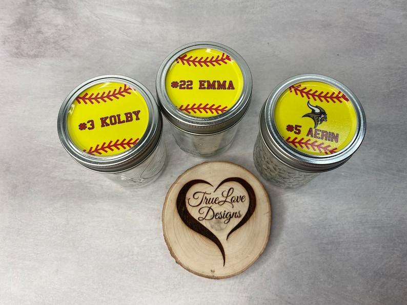 Puede incluir: Tres tarros de vidrio con tapas dise&ntilde;adas para parecerse a pelotas de softbol. Cada tapa tiene un fondo amarillo con costuras rojas y el nombre y n&uacute;mero de un jugador. Tambi&eacute;n hay un posavasos de madera en forma de coraz&oacute;n con el texto "True Love Designs".