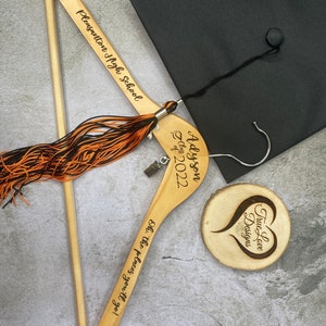 May include: A graduation-themed image featuring a black mortarboard cap, a wooden hanger with the text "Pleasanton High School", and a tassel. The hanger is engraved with "Adyson Class of 2022" and "Oh, the places you'll go!" A wooden coaster with a heart design is also present.