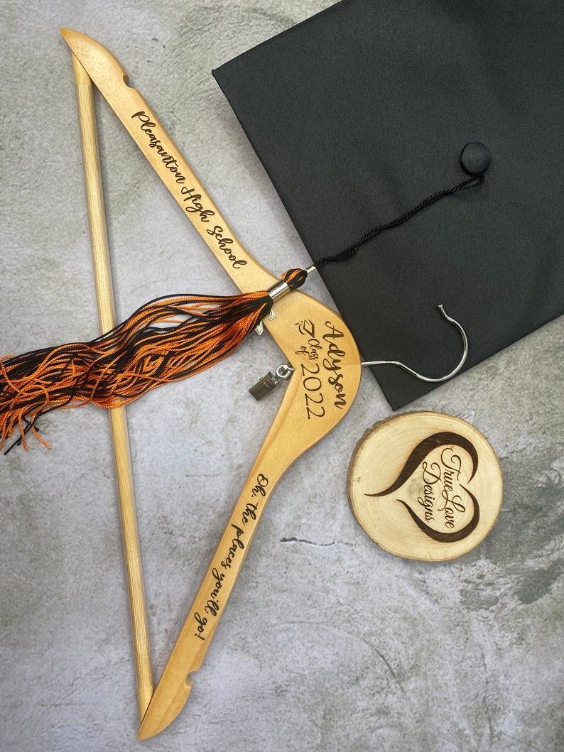 May include: A graduation-themed image featuring a black mortarboard cap, a wooden hanger with the text "Pleasanton High School", and a tassel. The hanger is engraved with "Adyson Class of 2022" and "Oh, the places you'll go!" A wooden coaster with a heart design is also present.