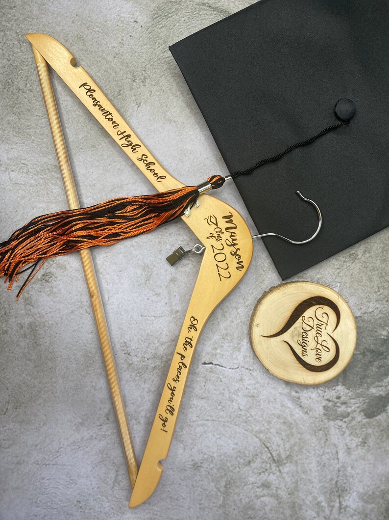 May include: A wooden graduation hanger with the text "Pleasanton High School" and "Oh, the places you'll go!" is displayed. A black graduation cap and a wooden coaster with a heart design are also visible. The hanger has a tassel with orange and black threads.