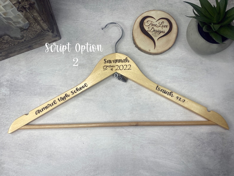 May include: A wooden clothes hanger with engraved text. The hanger has the words "Savannah Class of 2022", "Armorel High School", and "Isaiah 43:2". The hanger is light brown with a silver hook.