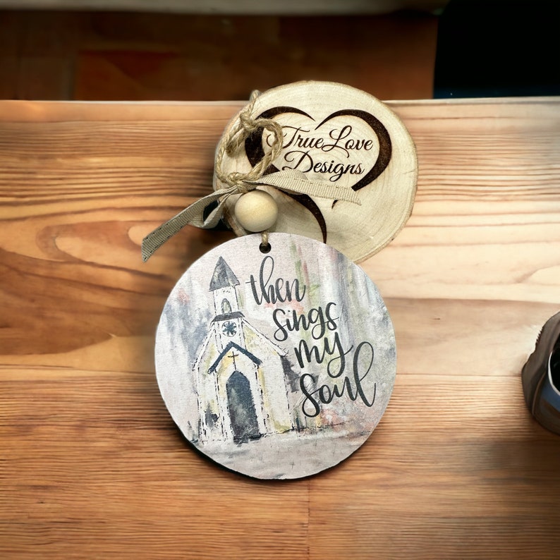May include: A round wooden sign with a watercolor painting of a church and the text "then sings my soul". The sign is attached to a smaller round wooden sign with a heart shape and the text "True Love Designs".