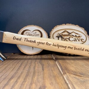 Puede incluir: Un martillo con mango de madera y cabeza de metal. El mango está grabado con el texto "Papá: Gracias por ayudarme a construir mi vida".