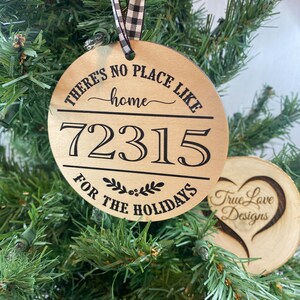 Op de afbeelding: Een ronde houten kerstversiering met de tekst "There's no place like home" en het nummer "72315" in zwarte letters. De versiering heeft ook de tekst "For the Holidays" en een klein kransontwerp. De versiering hangt aan een kerstboom.