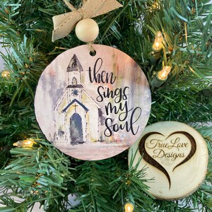 May include: A round wooden ornament with a watercolor painting of a church and the text "then sings my soul". The ornament is hanging on a Christmas tree. A smaller round wooden ornament with a heart shape and the text "True Love Designs" is also visible.