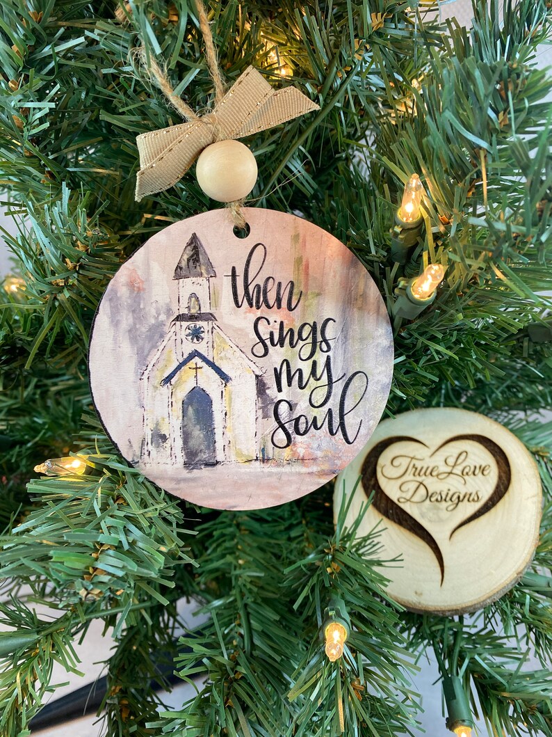 May include: A round wooden ornament with a watercolor painting of a church and the text "then sings my soul". The ornament is hanging on a Christmas tree. A smaller round wooden ornament with a heart shape and the text "True Love Designs" is also visible.