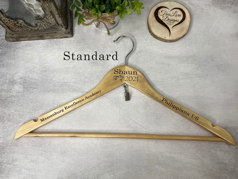 May include: A wooden clothes hanger with engraved text. The hanger has a metal hook and a small clip. Text includes "Shaun Class of 2021", "Massenburg Excellence Academy", and "Philippians 1:6". The hanger is light brown.