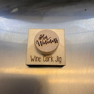 Pode incluir: Um jig de cortiça de vinho de madeira com o texto "Her Wine-ness" em uma rolha redonda. O jig é etiquetado como "Wine Cork Jig".