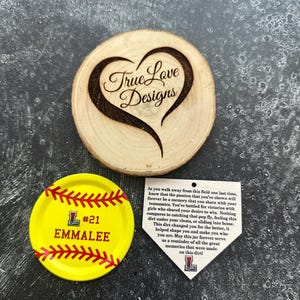 Puede incluir: Un adorno de madera en forma de coraz&oacute;n con "True Love Designs" grabado. Un posavasos amarillo con tem&aacute;tica de softbol con "#21 EMMALEE" y un dise&ntilde;o de costura de b&eacute;isbol. Una etiqueta blanca en forma de base con texto.