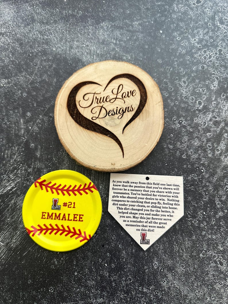Puede incluir: Un adorno de madera en forma de coraz&oacute;n con "True Love Designs" grabado. Un posavasos amarillo con tem&aacute;tica de softbol con "#21 EMMALEE" y un dise&ntilde;o de costura de b&eacute;isbol. Una etiqueta blanca en forma de base con texto.
