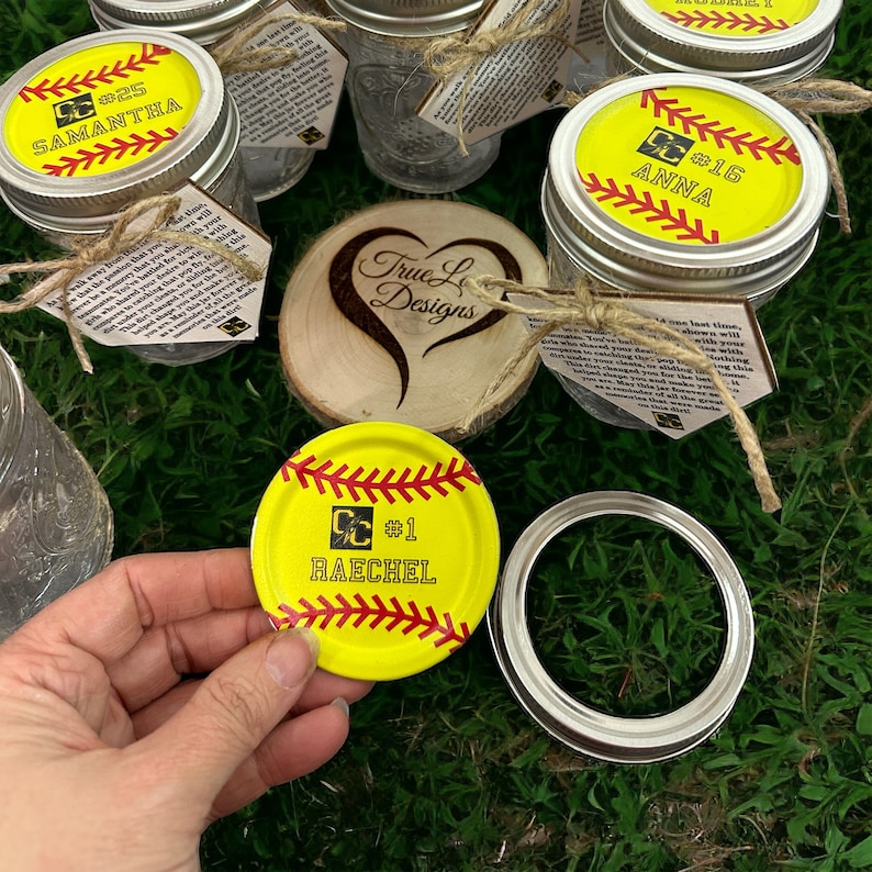 Puede incluir: Varios tarros de cristal con tapas amarillas con tem&aacute;tica de softbol. Las tapas presentan costuras rojas, nombres de jugadores y n&uacute;meros. Algunos tarros tienen etiquetas atadas con hilo. Tambi&eacute;n hay un coraz&oacute;n de madera con "True L Designs".