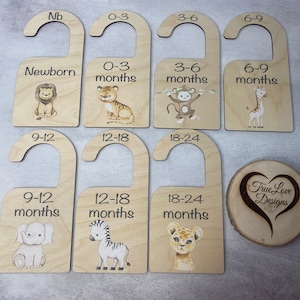 May include: Set of nine wooden hangers with animal illustrations and age ranges for baby clothes. The hangers are labeled with the following age ranges: Newborn, 0-3 months, 3-6 months, 6-9 months, 9-12 months, 12-18 months, 18-24 months. The animals include a lion, tiger, monkey, giraffe, elephant, zebra, and lion cub.